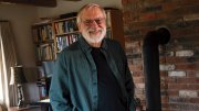 Tom Lee standing in his home in New Hampshire, surrounded by books and a brick fireplace.