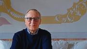 Werner Sollors seated on a couch with ornate wall designs in the background, wearing glasses and a dark sweater.