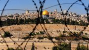 View of Jerusalem's Dome of the Rock framed by barbed wire, with the cityscape and hills in the distance.