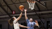 A Harvard player in white jumps toward the basket with the ball in her right hand, while a Columbia player in blue tries to block her shot 