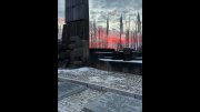 The Auschwitz-Birkenau memorial at sunset, with stone sculptures, commemorative plaques, and flags among bare trees.