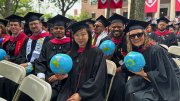 Graduates in caps and gowns hold inflatable globes at an outdoor ceremony with red banners in the background.