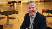 Tom Kane smiles  in a navy blazer and sits at a table in a modern room with empty chairs and tables behind him.