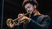 Toussaint Miller  in a dark sweater plays the trumpet on stage with eyes closed.