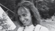 A black-and-white portrait of a smiling person with wavy hair, wearing a beaded necklace.