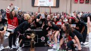 Players shouting with joy surround a large trophy while a fan in the background holds up a giant replica ticket to the NCAA tournament.