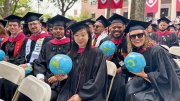 Graduates in caps and gowns smile while holding inflatable globes during an outdoor commencement ceremony.