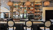 Elegant bar with glass shelves stocked with liquor bottles, three cane-back chairs, and hanging pendant lights.