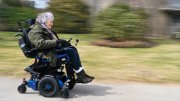 Lisa Iezzoni smiling while riding a motorized wheelchair on a paved path through a grassy area