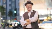 Salvador Litvak smiles, arms crossed, in a vest and hat on a city street.