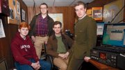 Four students in a wood-paneled radio room with monitors and radio equipment, one wearing a Harvard sweatshirt.
