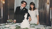 A bride and groom in formal attire cut a tiered cake at a candlelit wedding reception table.