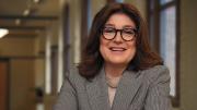 Nicolo Maestas in a grey suit and wearing glasses sits with her arms on a table