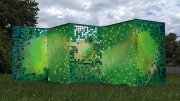 A six-foot-tall, five-panel folded screen stands in a field of grass next to the woods. It's painted different shades of green, with some squares cut out that represent digital pixels.
