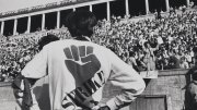 Black-and-white photo of a person wearing a “STRIKE” shirt facing a large crowd in a stadium during a protest.