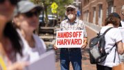Man, standing in small group of people outside the courthouse, holding a sign that reads "HANDS OFF HARVARD" in red letters
