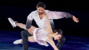 Figure skating pair performs an elegant lift, with the woman extended low and arm reaching toward the ice.