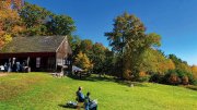 People sit in lawn chairs near a rustic barn at Cider Garden in New Salem on a sunny day.