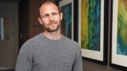Man in gray sweater standing in hallway with colorful abstract art on wall.