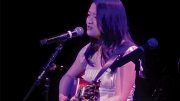 Julie Riew, wearing a white dress, playing guitar and singing into a microphone on stage.