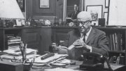 David McCord in suit reading a book at cluttered wooden desk in office filled with framed art and shelves.