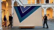 Two people moving large abstract painting with blue V-shaped design in museum courtyard.