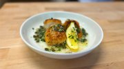 Golden-brown scallops garnished with herbs and lemon in a white bowl on a wooden surface.