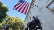 Bronze statue of John Harvard  with an American flag in the background, surrounded by trees.