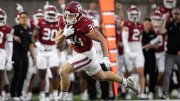 A football player in a maroon jersey sprints past teammates on the sideline.