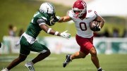 A football player in red dodges a tackle from a defender in green on a grassy field.
