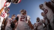 Harvard football team huddles on the field under a clear blue sky before the game.