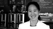 A smiling Joanne Chang in a white coat stands behind a display of pastries in Flour Bakery.