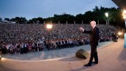 A speaker addresses a large crowd at an outdoor evening event.