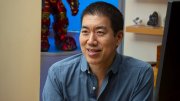 Professor David Liu smiles while sitting at a desk with colorful lanterns and a figurine in the background.