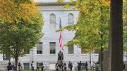 People gather near the John Harvard Statue in front of University Hall surrounded by autumn trees.