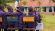 Students in purple jackets seated on chairs, facing away in a grassy area.