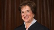 Justice Elena Kagan in a black robe, smiling against a wooden panel background.