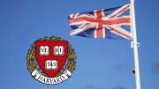 Union Jack flag waving next to the Harvard University crest against a blue sky.
