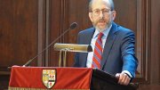 A man in a suit speaking at a podium with a red drape and a university crest.