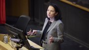 A woman in a gray suit gestures while speaking at a podium in a lecture hall.