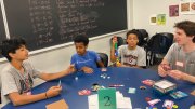 Four young people sitting around a table playing a card game, with a chalkboard in the background.