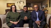 Nancy Hill, Rendall Howell, and their son Theo sit together on a couch in a cozy living room, smiling at the camera.