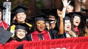 Graduates celebrate joyfully, wearing caps and gowns, with some waving and smiling.