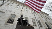 A seated statue of a man reading, with an American flag waving in the background.