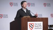 A speaker gestures while presenting at a podium with Harvard Radcliffe Institute branding.