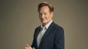 Conan O’Brien in a dark suit and light shirt, smiling and posing with hands clasped.
