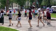 A group of people march in a park holding signs, surrounded by greenery and trees.