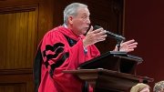 Former Harvard President Lawrence S. Bacow in a red academic robe gestures passionately at a podium, with seated audience members behind.