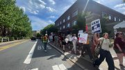 Protesters holding signs march along a sunny street in front of a building.