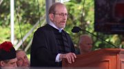 Harvard President Alan Garber in academic robes speaks at a podium outdoors, with a blurred audience behind him.